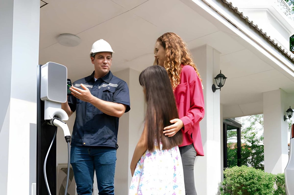 Technician in hard hat installing electric vehicle charging station at home entrance while woman and child observe