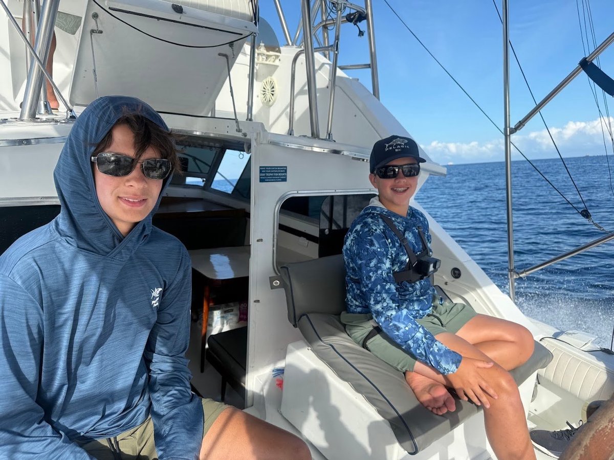 Two people on a fishing boat in blue water, one wearing a blue hoodie and sunglasses on the left, another in a blue patterned shirt on the right