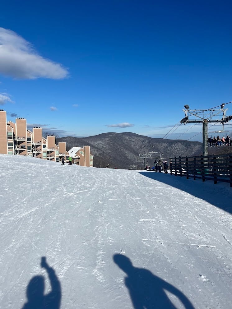 Snowy ski resort with lodge buildings, ski lift, mountains in background, and two peoples shadows on white slope under blue sky