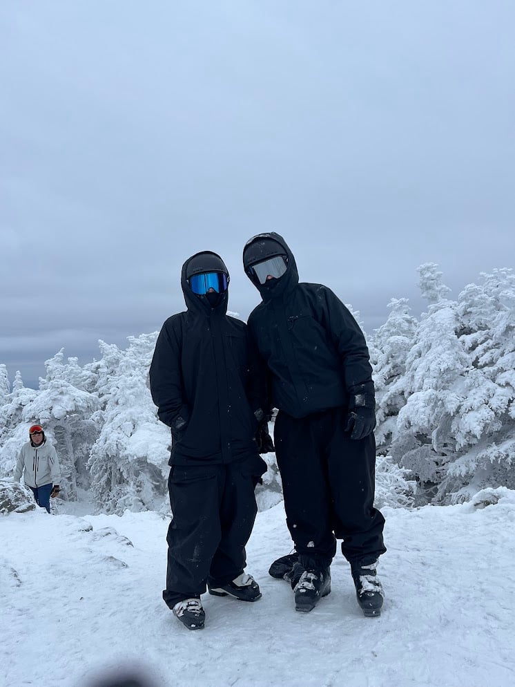 Two skiers in black jackets and helmets posing on snowy terrain with snow-covered trees in background