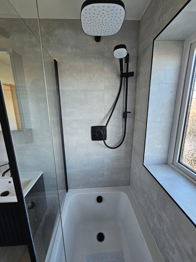 Modern bathroom shower with rainfall showerhead, handheld spray, white bathtub, gray tile walls, glass enclosure, and window