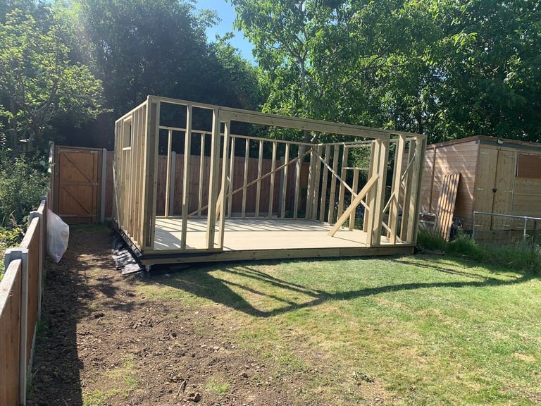 Wooden shed frame under construction in a fenced backyard with green grass and trees in the background
