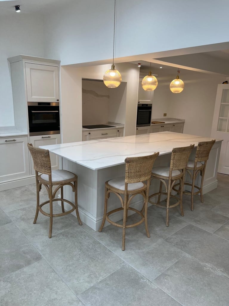Modern kitchen interior with white cabinetry, large island with seating, rattan bar stools, pendant lighting, and embedded appliances