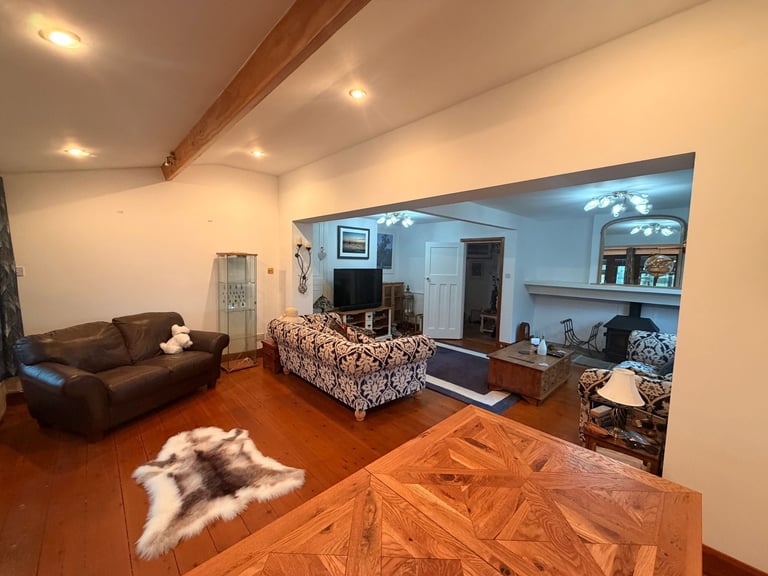 Open-concept loft living room with brown leather furniture, wooden floors, exposed beam, and adjoining kitchen area with white walls and recessed lighting