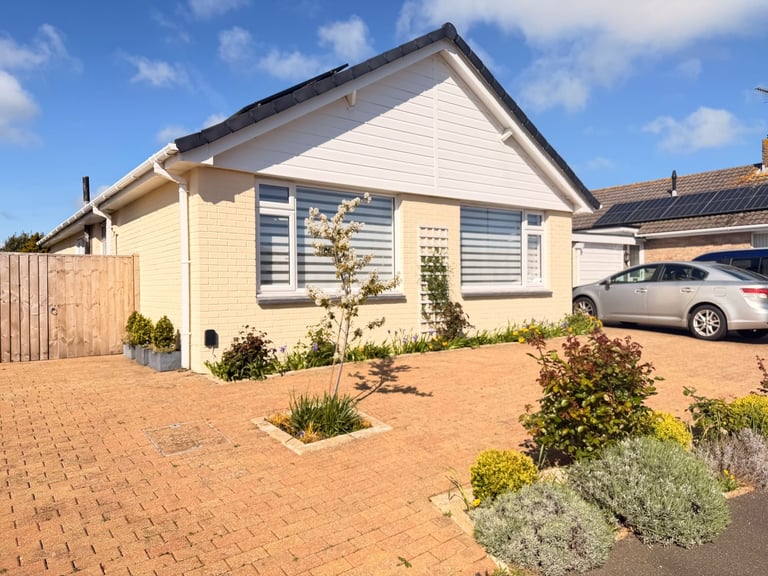 Single-story cream-colored house with gray roof, shuttered windows, brick paved driveway with landscaping, and parked cars visible in the background