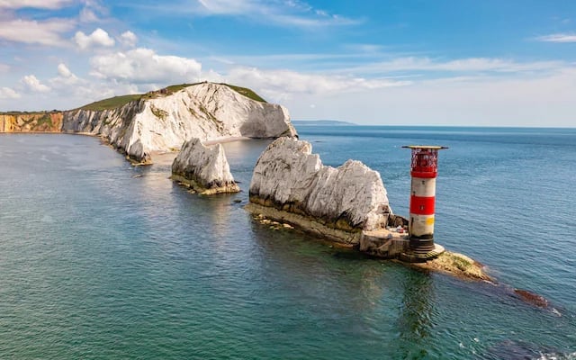 Red and white striped lighthouse standing near white chalk sea stacks in turquoise ocean water under blue sky