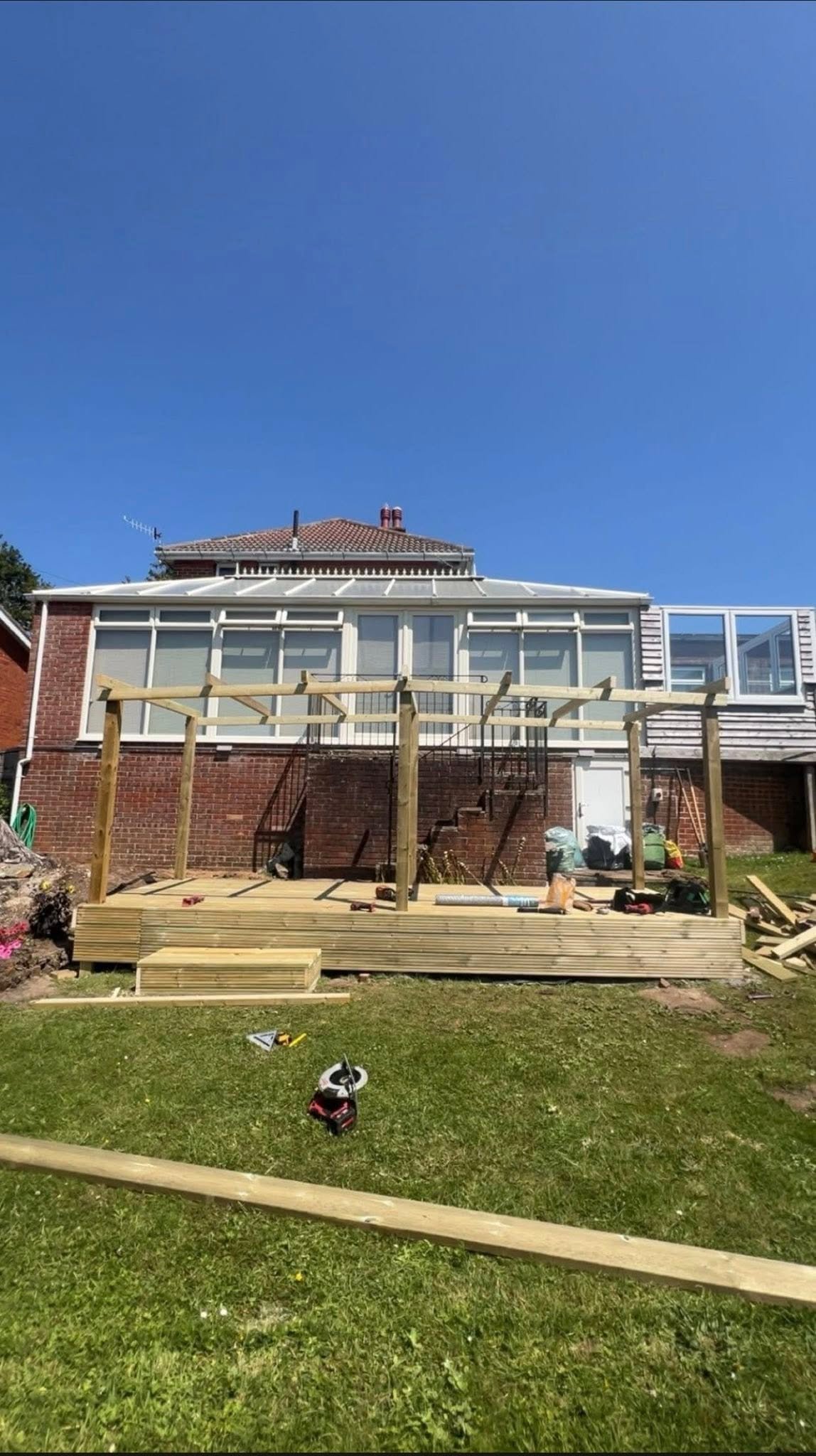 Wooden deck and pergola structure attached to brick house with green lawn and clear blue sky