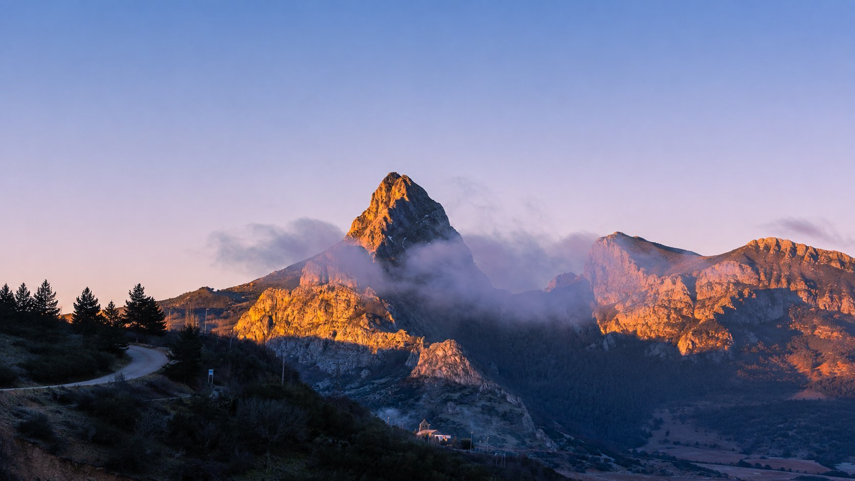 Mountain range at sunrise with dramatic rocky peaks illuminated by golden light, mist in valleys, evergreen trees in foreground