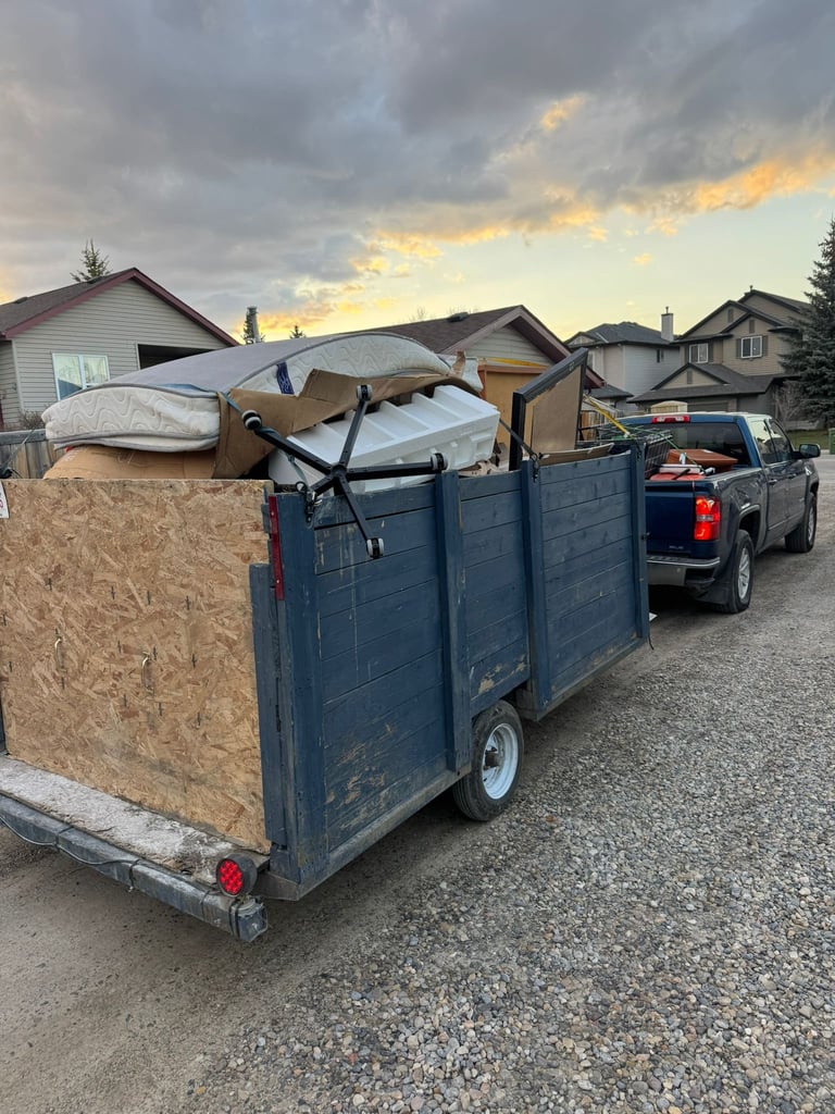 Loaded blue trailer attached to pickup truck carrying furniture and cardboard boxes on residential street at dusk