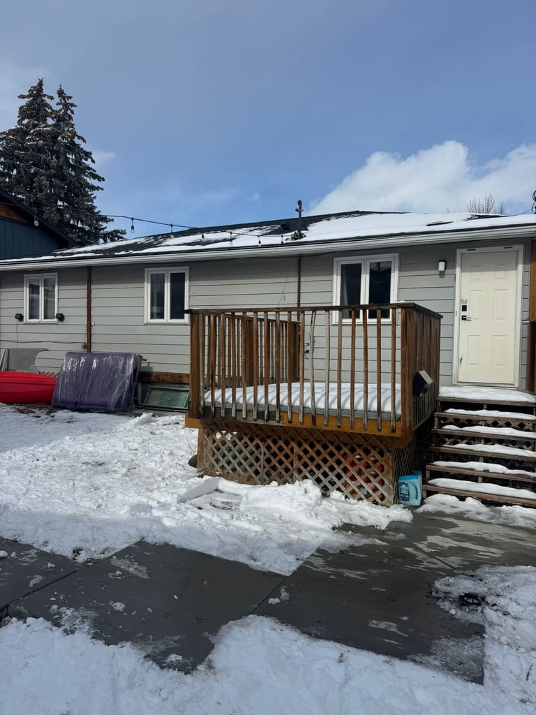 Single-story beige house with wooden deck and railings in winter, surrounded by snow and bare ground