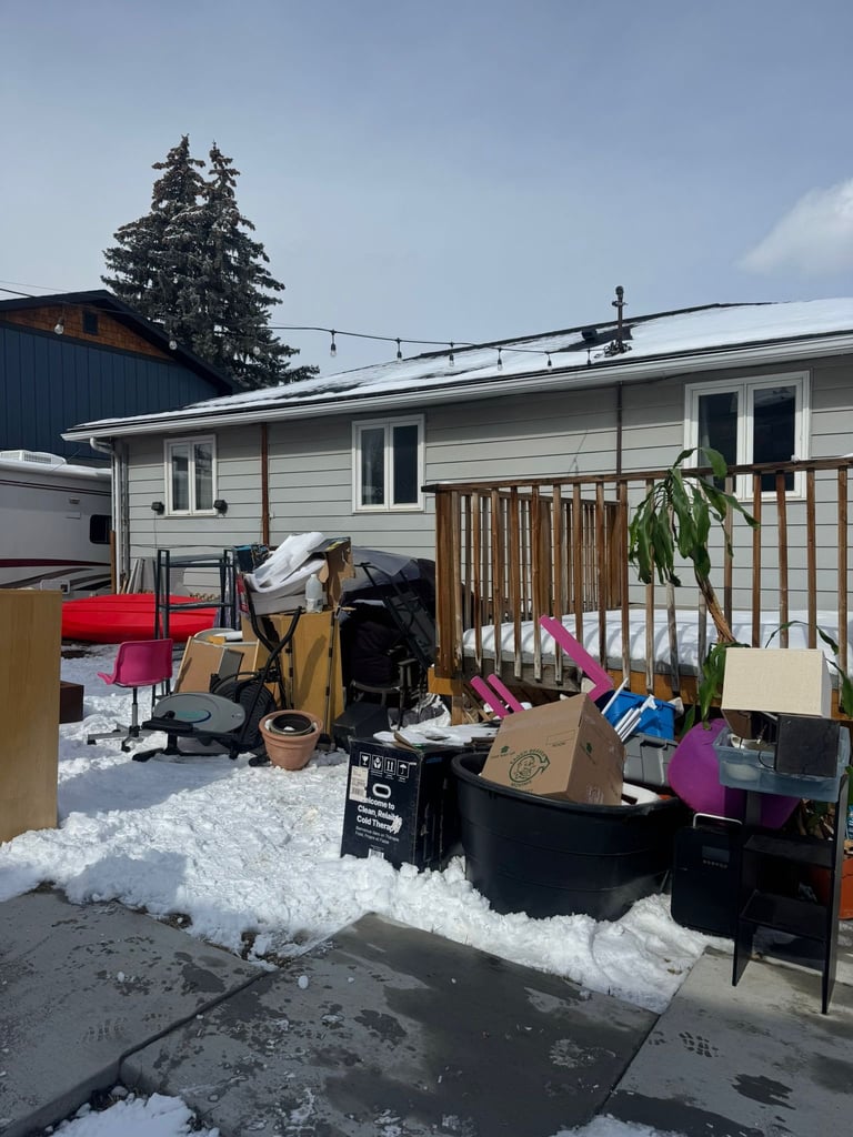 A cluttered front yard and patio of a mobile home in winter, with scattered boxes, furniture, and belongings on snow-covered ground.