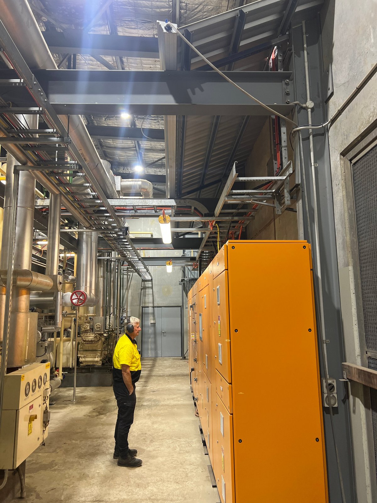 Worker in yellow safety vest standing in industrial facility corridor lined with orange equipment and machinery