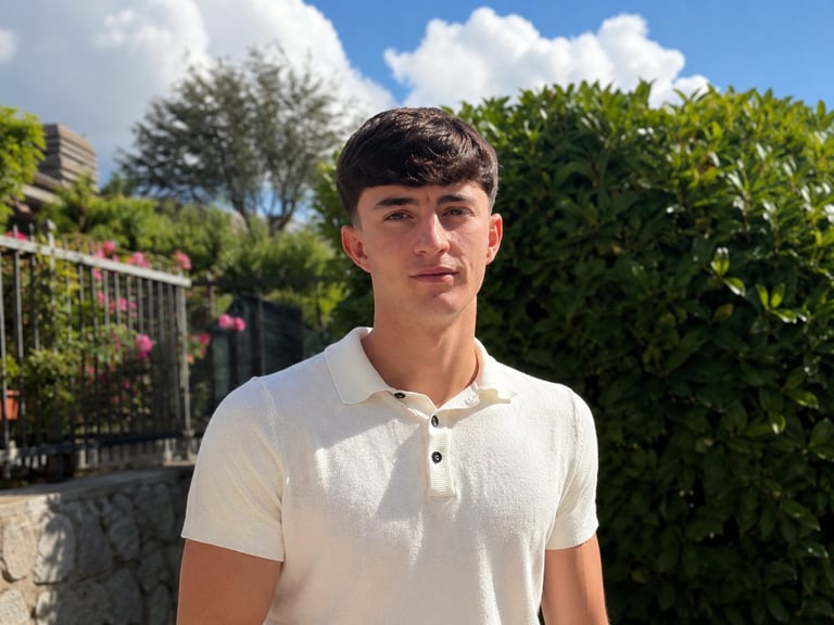 Young man wearing a white polo shirt standing outdoors in a garden setting with green hedges and fence behind him