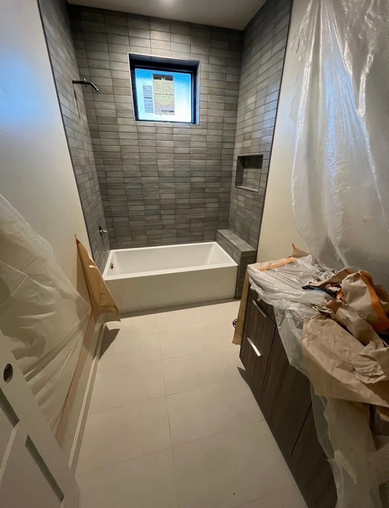 Modern bathroom under renovation with gray tile walls, white soaking tub, and construction materials covered with plastic sheeting