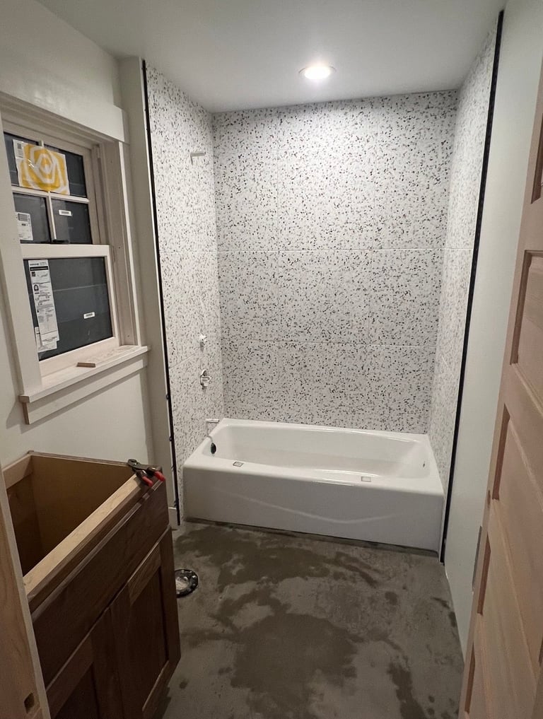Modern bathroom under renovation with white speckled tile surround, acrylic bathtub, recessed lighting, and wooden vanity cabinet