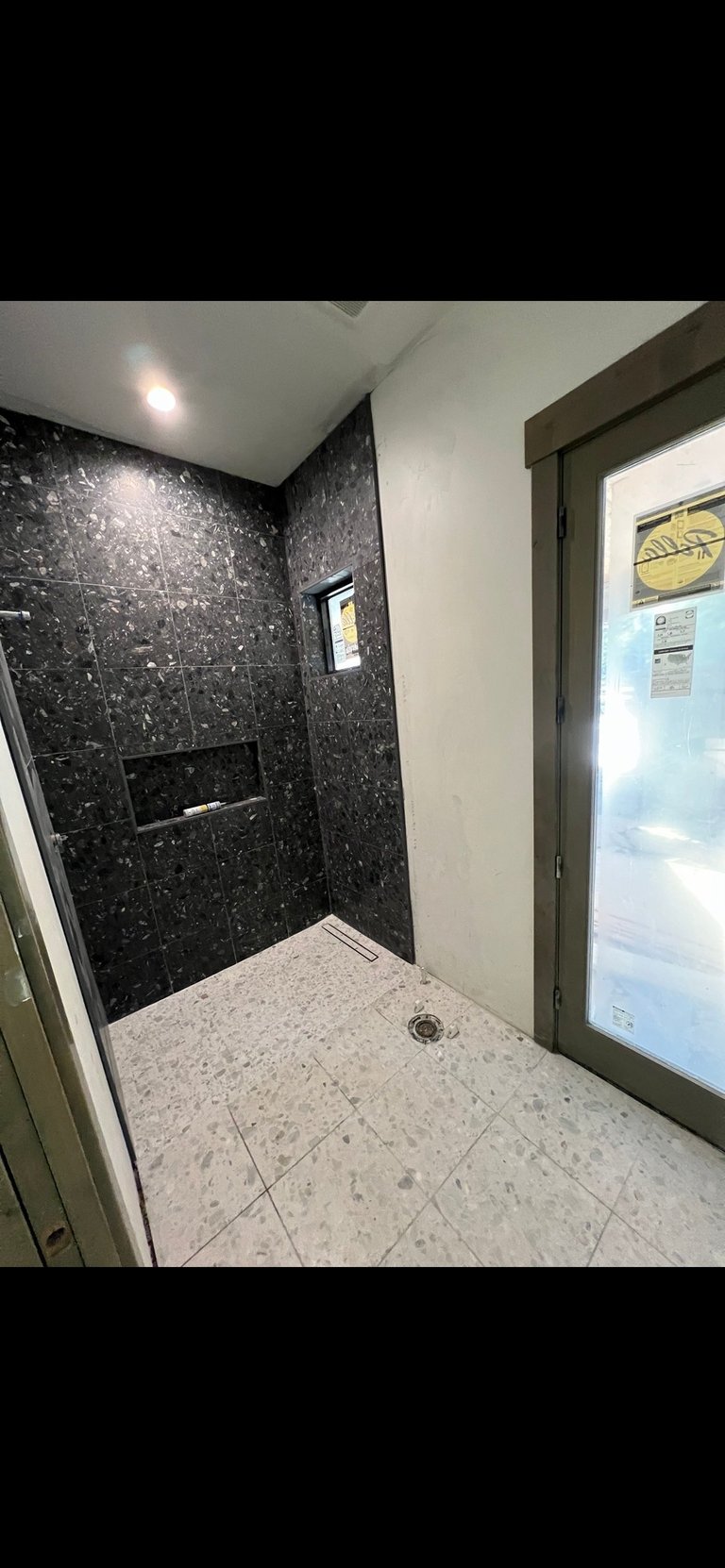 Modern bathroom with black marble accent wall, white speckled floor tiles, recessed lighting, and glass shower enclosure