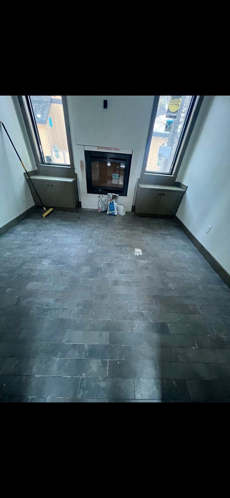 Empty modern apartment interior with dark tile flooring, fireplace, large windows overlooking city buildings, and minimal furnishings