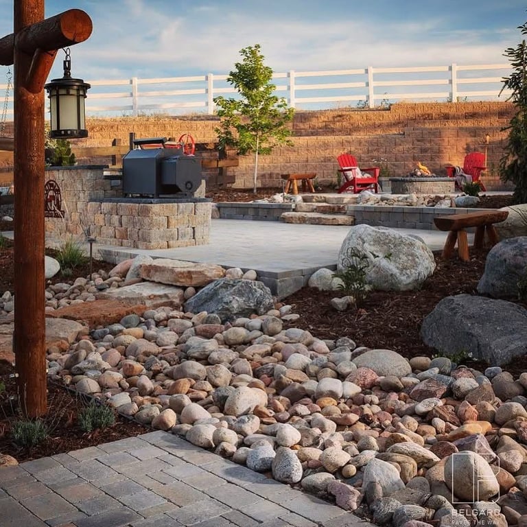 Outdoor patio with stone fireplace, red Adirondack chairs, river rock landscaping, and scenic overlook of distant cliffs at sunset