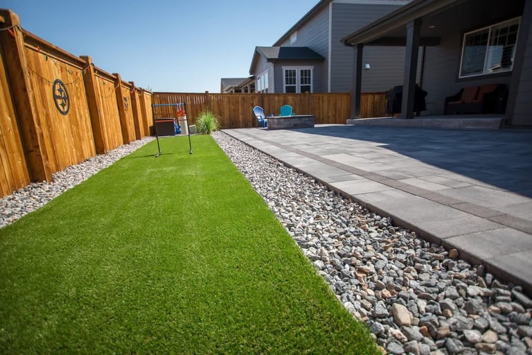 Modern backyard with green lawn bordered by white gravel, paved patio area, and wooden fence with house in background