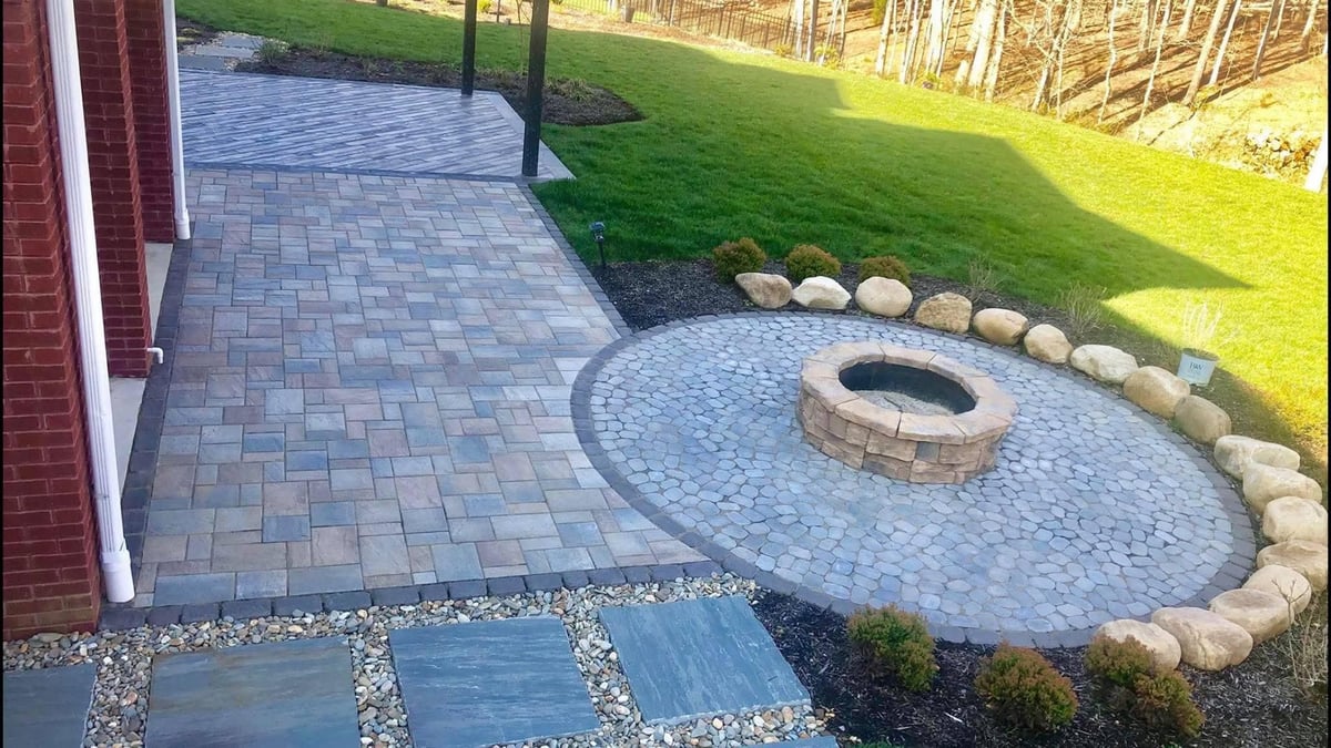 Backyard patio with gray paver walkway, fire pit ringed by boulders, green lawn, and wooden fence