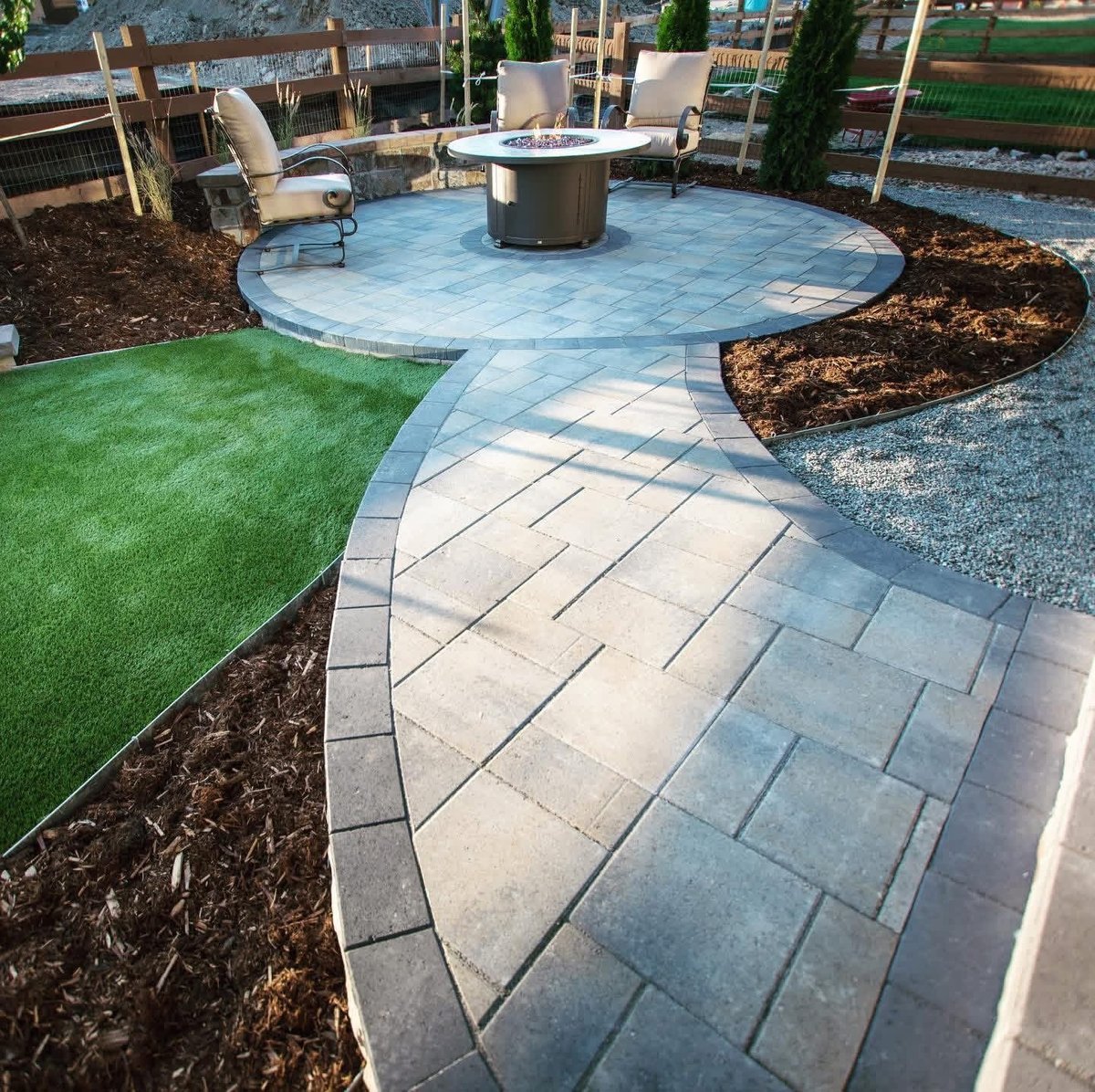 Backyard patio with circular fire pit, curved pavers, lounge chairs, artificial turf, mulch beds, and metal fencing