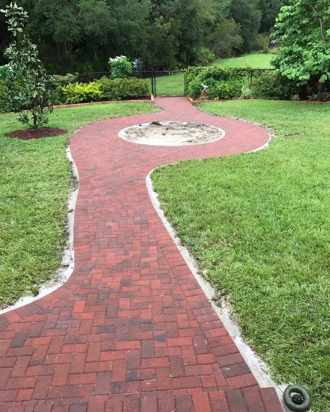 Curved red brick pathway through green lawn leading to round patio with fire pit, bordered by trees and fencing