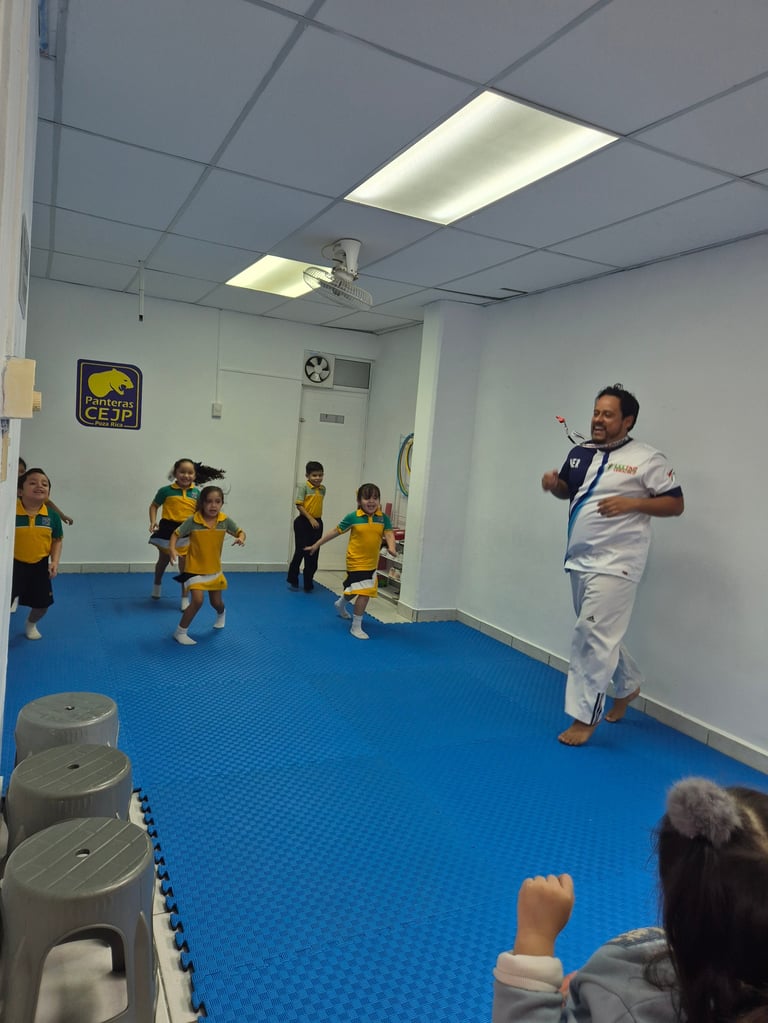Instructor in white uniform demonstrates martial arts to young children in yellow shirts in an indoor gym with blue flooring