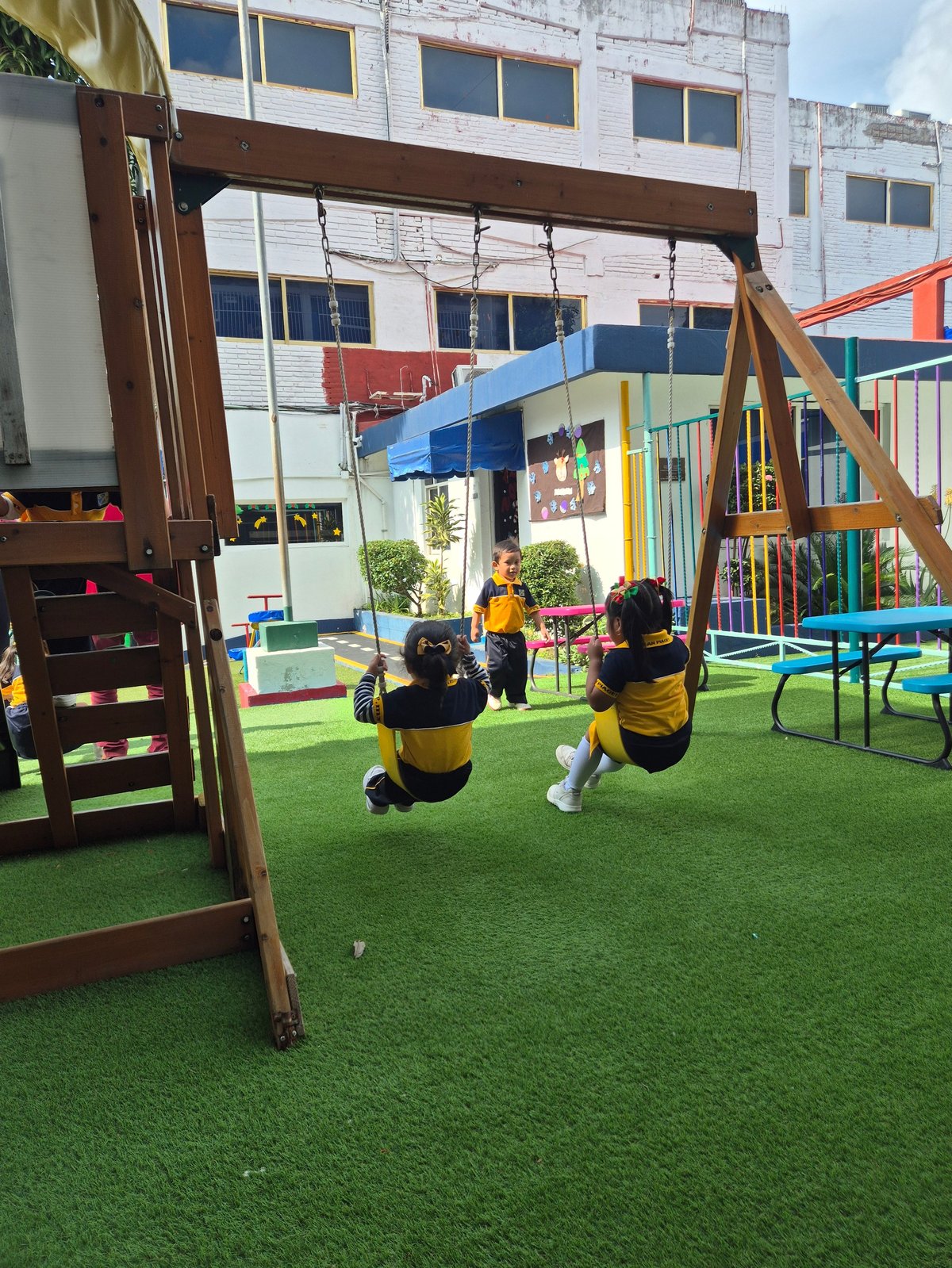 Children playing on swings in a colorful outdoor school playground with artificial grass and modern building in background