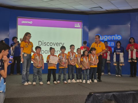 Group of children in yellow and orange shirts standing on stage at an awards ceremony receiving recognition.