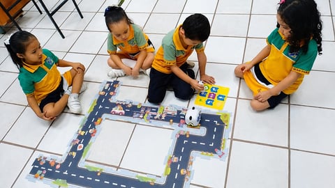 Four children in yellow and green uniforms sitting on a tile floor playing with a toy road mat and vehicles.