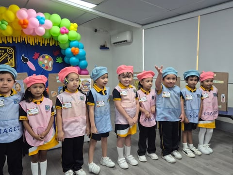 Group of children wearing pink and blue aprons and hats standing in a classroom decorated with colorful balloons.