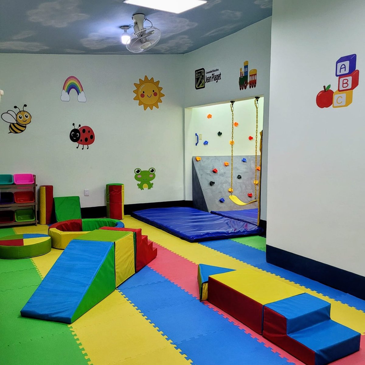 Colorful indoor playroom with padded foam play structures, climbing wall, and wall decorations featuring rainbows, sun, and insects.
