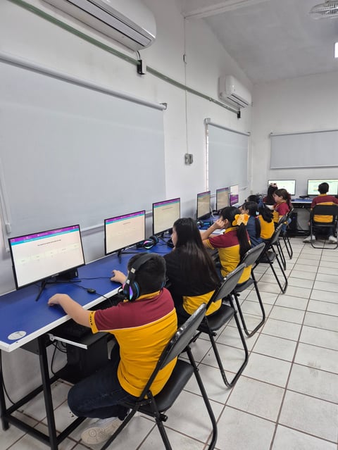 Students in yellow vests working at computers in a classroom with blue tables and whiteboards