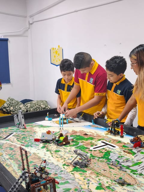 A group of young students in yellow and navy uniforms work together on a robotics project with LEGO models and building materials on a table in a classroom.