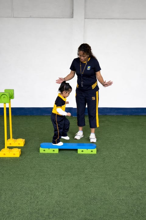 Adult instructor in navy and yellow sportswear guides a small child on a balance beam during a training session on artificial turf.