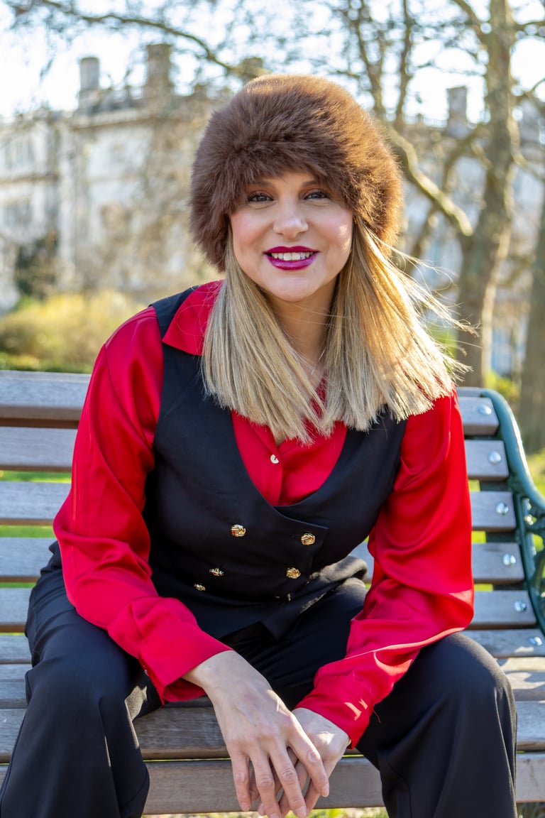 Woman sitting on a bench wearing a red and black blazer and brown fur hat, smiling at camera with a historic building in the background