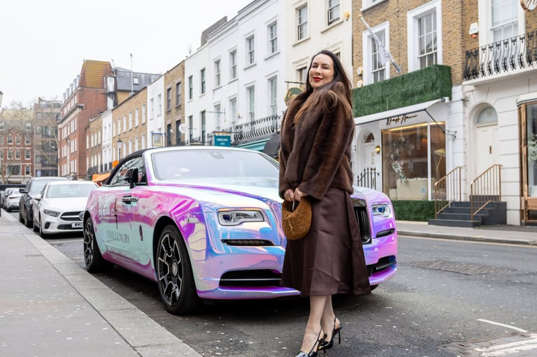 Woman in brown coat standing beside pink and blue luxury car on London street with historic buildings