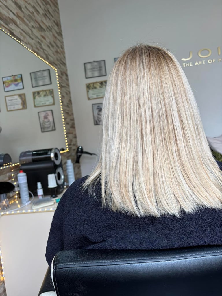 Back view of woman with long straight platinum blonde hair sitting in salon chair facing illuminated mirror with products and certifications displayed
