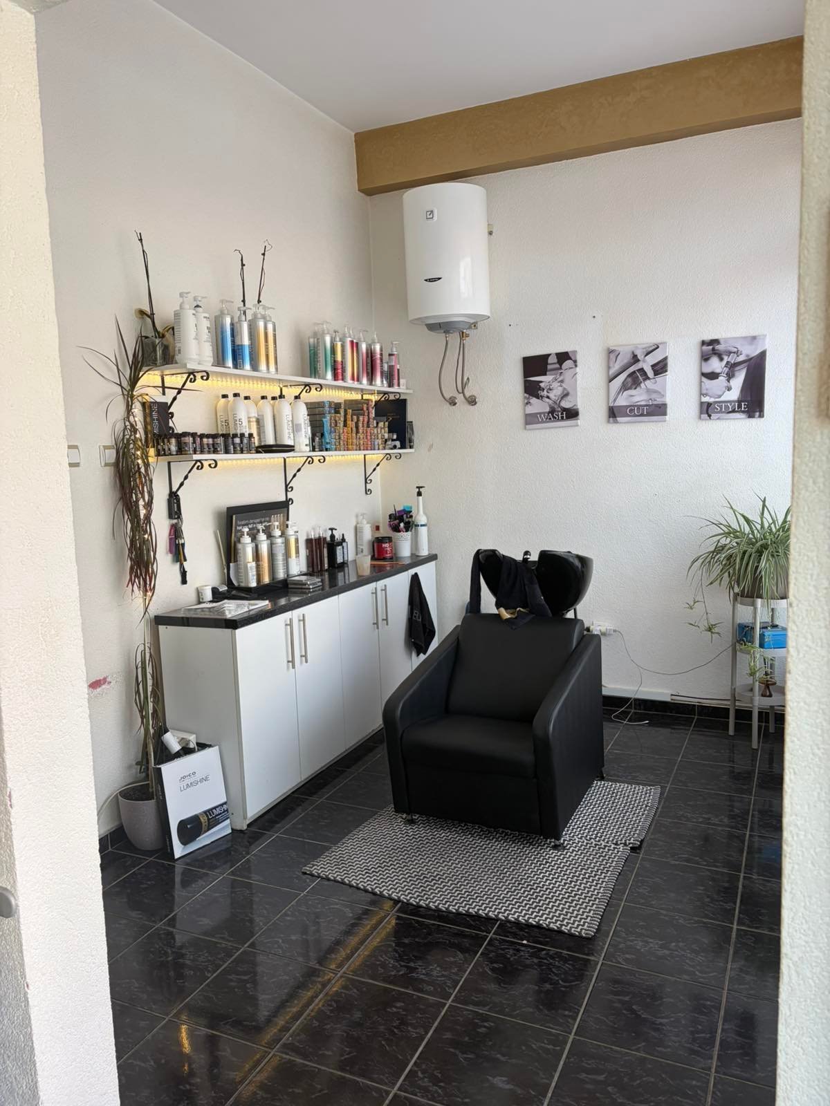 Interior of a modern salon or beauty studio with black tile flooring, white cabinetry with shelves of products, a black styling chair, and wooden beam ceiling detail.