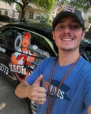 Man in blue shirt and cap giving thumbs up gesture next to a black vehicle with racing graphics and signage