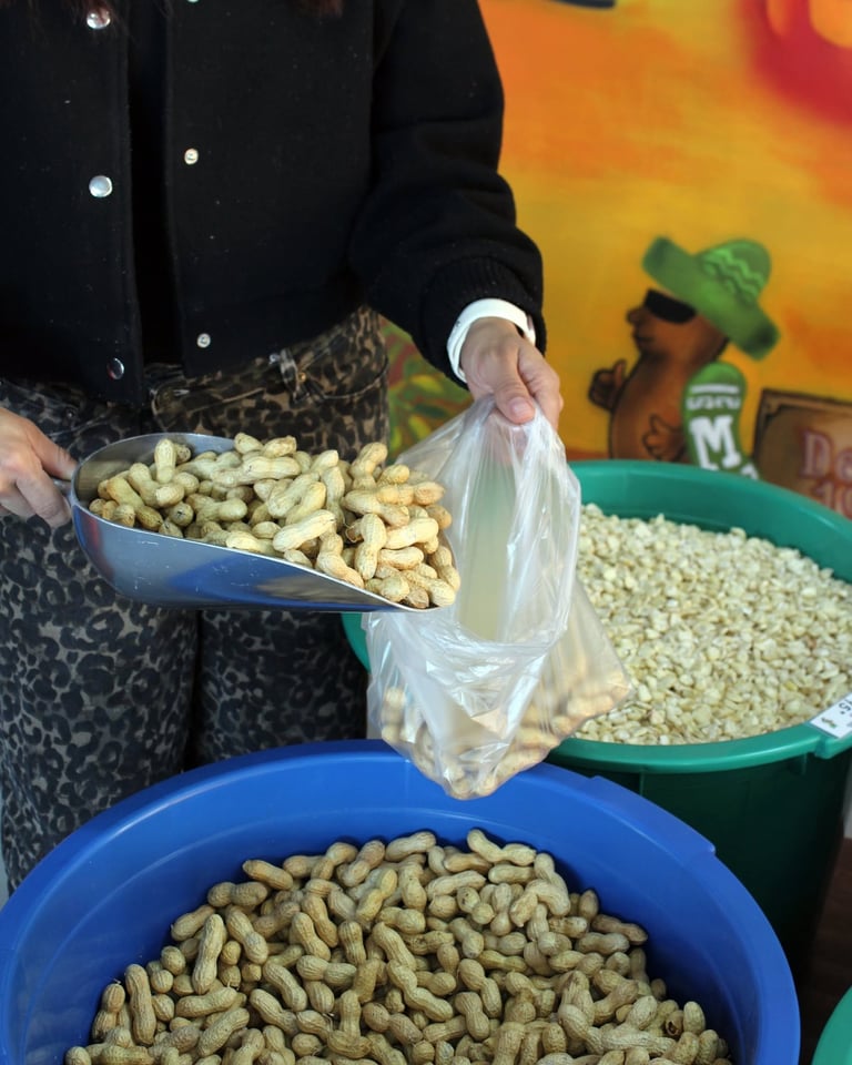 Vendor scooping roasted peanuts into a plastic bag at outdoor market stall with colorful signage