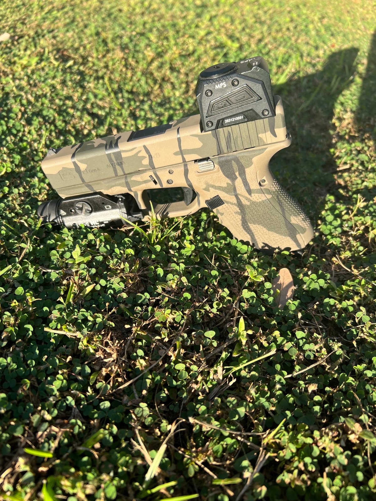 Tan and black tactical pistol with mounted red dot sight lying on green grass outdoors with shadow