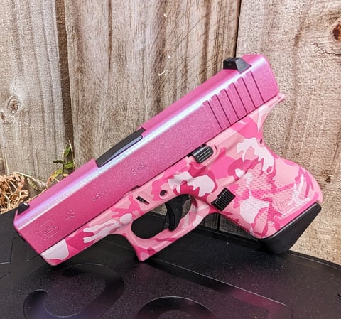 Pink and white camouflage handgun with black accents displayed on a wooden background