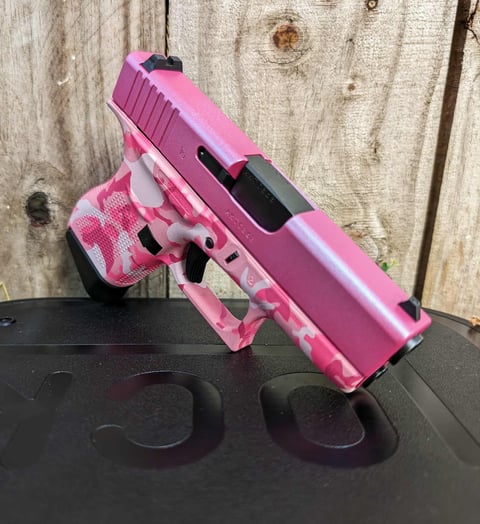Pink and camo tactical handgun with black accents sitting on a black surface against a weathered wooden wall background