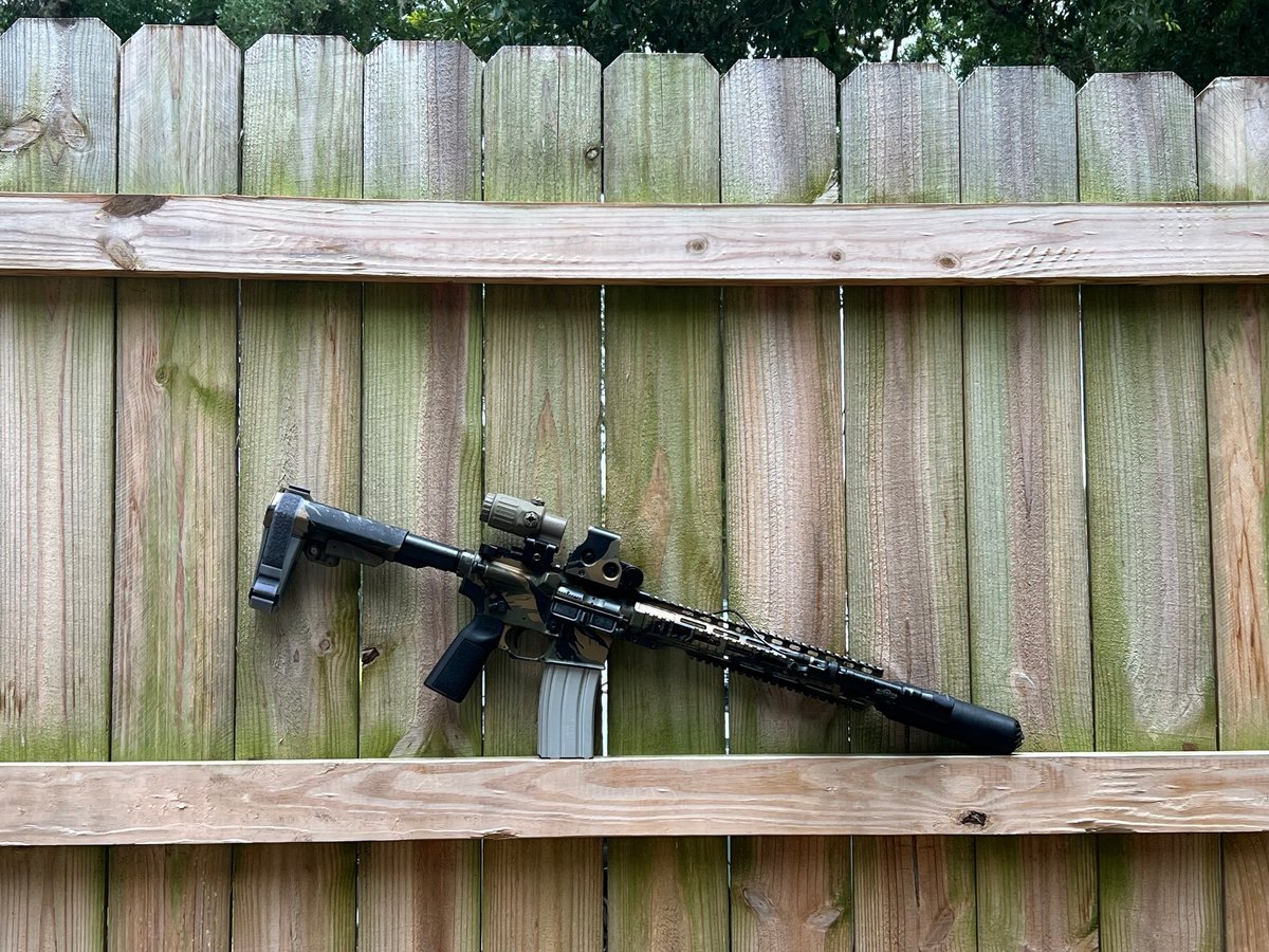 Black tactical rifle with scope mounted on wooden shelf against weathered wooden fence background