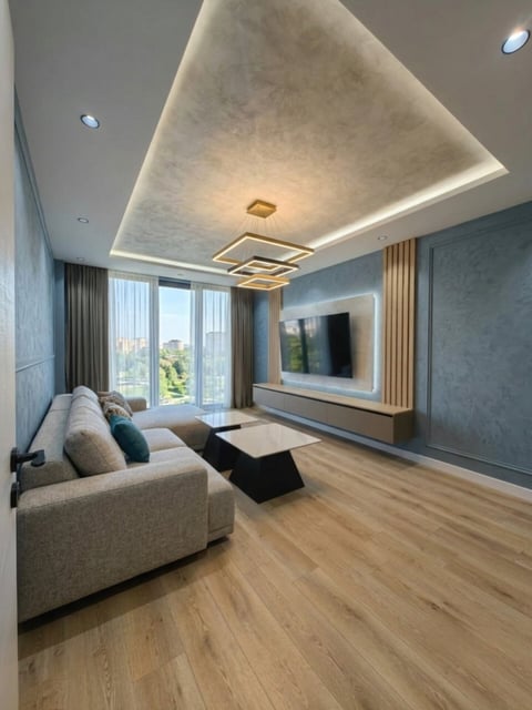 Modern living room with beige sofa, wall-mounted TV, coffered ceiling with accent lighting, wooden flooring, and floor-to-ceiling windows