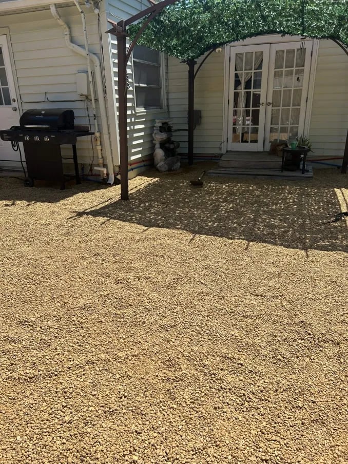 Backyard patio with gravel surface, pergola, grill, and RV garage entrance with white doors and siding.