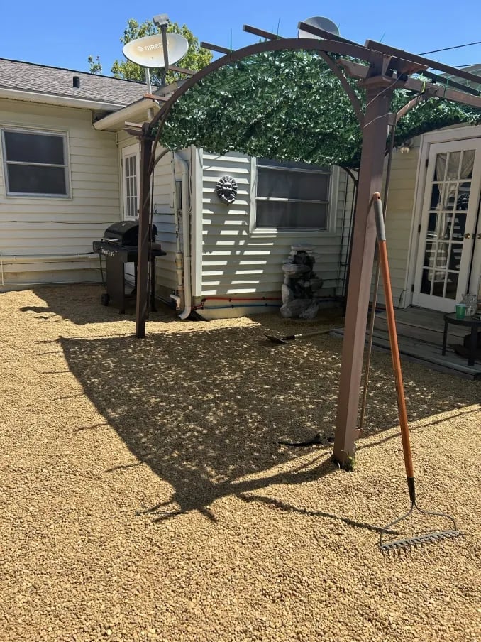 A wooden pergola with climbing ivy covers a shaded seating area in a gravel backyard next to a house