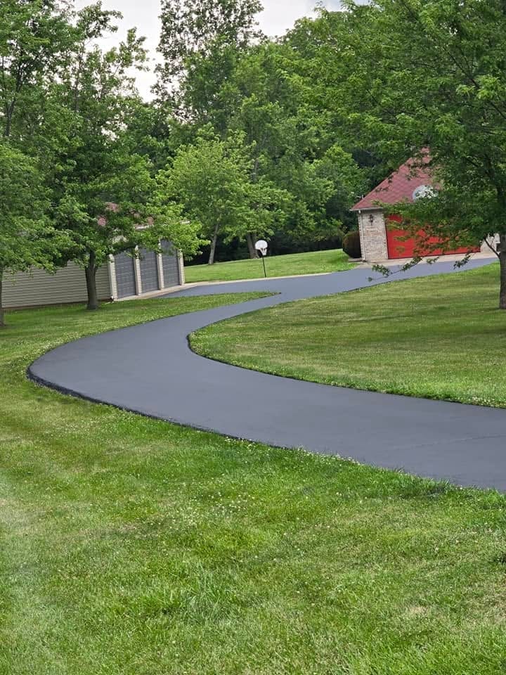 Curved asphalt driveway winding through a manicured lawn with mature trees and a brick building with red roof in the background