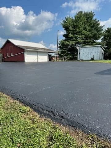 Rural farm with red barn, white garage, and gray storage shed on asphalt driveway under blue sky with clouds